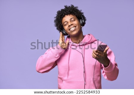Happy gen z African American guy wearing headphones holding mobile using cellphone dancing enjoying listening online streaming music on cell phone, standing isolated on light purple background. Royalty-Free Stock Photo #2252584933