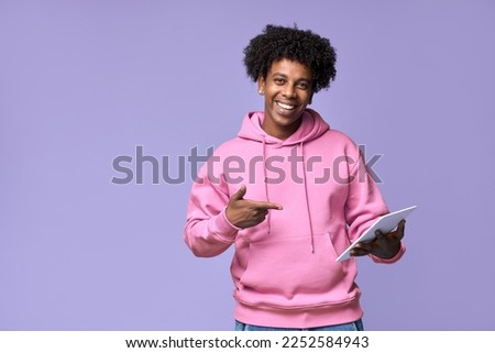 Happy cool African American teenage guy teen boy model wearing pink hoodie holding tab using digital tablet pointing at device advertising online learning isolated on light purple background. Royalty-Free Stock Photo #2252584943