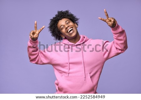 Young cheerful happy hipster African American teen guy wearing pink hoodie isolated on light purple background. Smiling cool joyful ethnic generation z teenager model laughing showing peace sign.