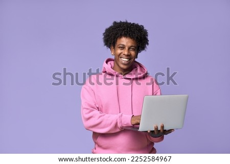 Young happy African American teenager student boy wearing pink hoodie holding laptop using computer technology advertising elearning education and online webinars isolated on light purple background.
