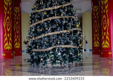 A Christmas tree with whitish green leaves with golden hanging ornaments and circled by a golden yellow ribbon.