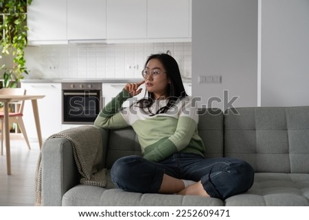 Concentrated pensive Asian girl student looks away and touches face with hand when planning trip to college. Carried away chinese woman sits on sofa in apartment thinking about university education