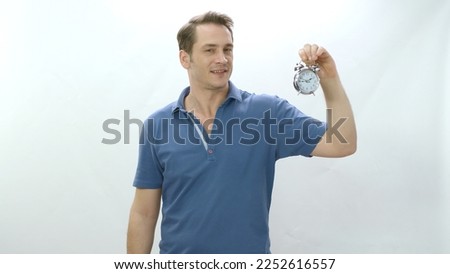 Young man holding a small clock isolated on a white background. The man explains that time is important and that it is running out. He shows the small desk clock to the camera.