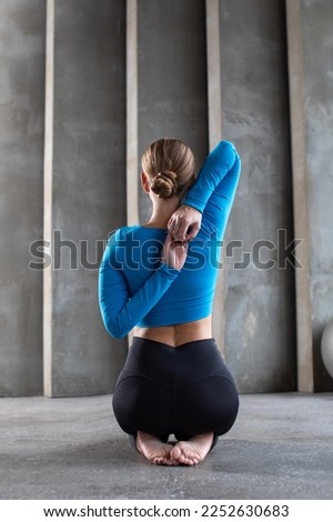 A young woman in sportswear sits with her back and locks her hands behind her back, stretching the shoulder muscles.  Royalty-Free Stock Photo #2252630683