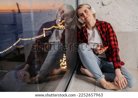 Blonde American girl in casual plaid shirt and blue jeans sitting on windowsill using headphones, phone listening music enjoys sunset and city lights being in romantic moos. Dreamer, woman in love.