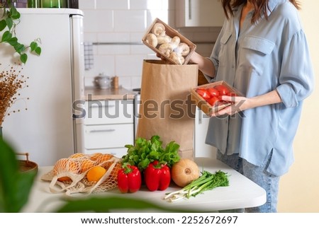 Woman at home getting groceries out of a shopping bag with grocery ordered from internet. Fresh organic vegetables, greens and fruits. Kitchen interior.  Food delivery concept Royalty-Free Stock Photo #2252672697