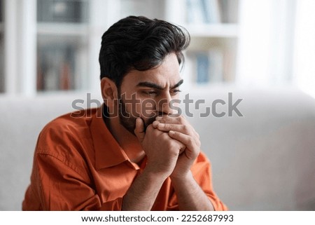 Closeup photo of sad depressed attractive bearded young middle eastern man in orange shirt sitting on couch at home, covering his mouth with hands, thinking about something, copy space Royalty-Free Stock Photo #2252687993
