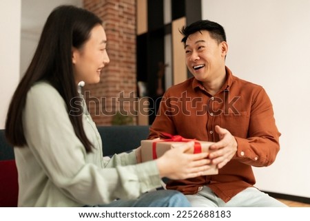 Loving young asian wife giving present to her happy mature husband, sitting on couch at home. Positive woman greeting spouse with birthday or anniversary, surprising with gift box