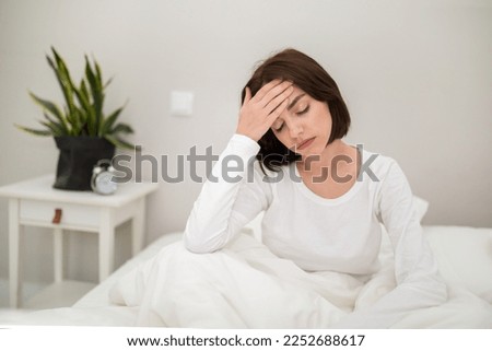 Tired unhappy young beautiful brunette woman waking up with headache in the morning, sick lady sitting in bed at home, touching her head, suffering from hangover or migraine, copy space Royalty-Free Stock Photo #2252688617