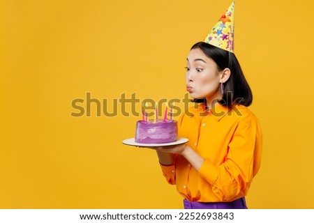 SIde view happy fun young woman wear casual clothes hat celebrate hold purple cake blow out candles make wish in special moment isolated on plain yellow background. Birthday 8 14 holiday party concept