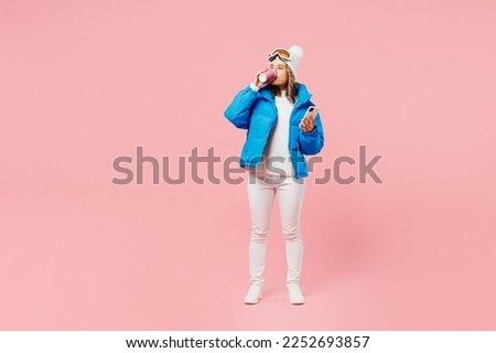 Snowboarder woman wear blue suit goggles mask hat ski padded jacket use hold mobile cell phone drink sip coffee isolated on plain pink background. Winter extreme sport hobby weekend trip relax concept