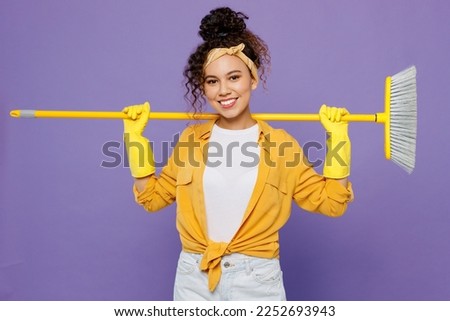 Young fun smiling happy housekeeper woman wear yellow shirt tidy up hold broom behind herself sweeps floor clean house isolated on plain pastel purple background studio. Housework housekeeping concept Royalty-Free Stock Photo #2252693943