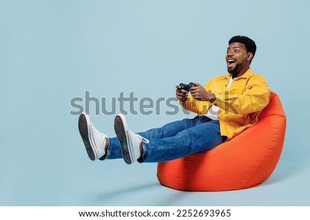 Full body young man of African American ethnicity 20s wear yellow shirt sit in bag chair hold in hand play pc game with joystick console isolated on plain pastel light blue background studio portrait.