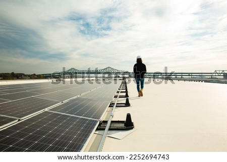 Female solar installer inspecting a commercial rooftop solar system in Charlestown, Boston, Massachusetts, United States with the Tobin Bridge in the background.