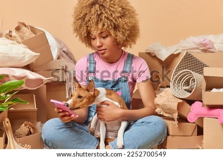 Serious curly woman checks notification on smartphone focused at screen poses in lotus pose with dog searches new apartment to rent packs personal belongings in carton boxes. Real estate concept