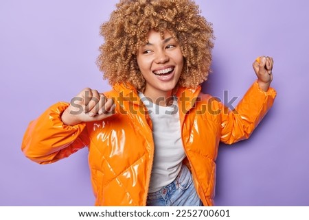Curly haired woman smiles broadly shows white teeth dances carefree has playful expression wears fashionable orange jacket has happy mood isolated over purple background. Cheerful female dancer
