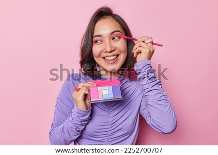 Happy Asian woman with dark hair applies pink eyeshadow uses cosmetic brush smiles broadly has white even teeth wears purple turtleneck isolated over pink background. Beauty and wellness concept
