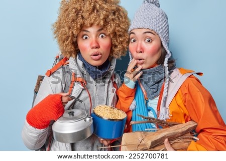 Photo of two frightened women stare impressed reacts to something breathtaking have picnic together hold kettle pot with noodles carry wood for making fire dressed in outerwear isolated on blue wall