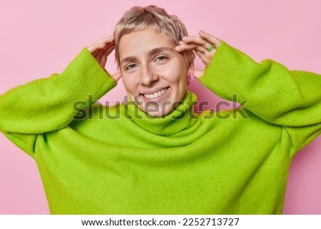 Beautiful cheerful woman with short haircut keeps hands on head smiles broadly has dimples on cheeks white even teeth wears loose green jumper looks happily at camera isolated over pink background Royalty-Free Stock Photo #2252713727
