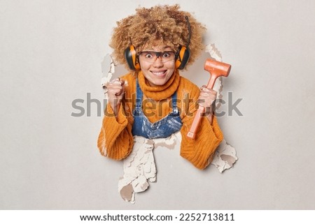 Curly haired female builder holds hammer clenches fist maintainces house renovation service wears glasses headphones on ears dressed in orange jumper and overalls breaks through plaster wall Royalty-Free Stock Photo #2252713811