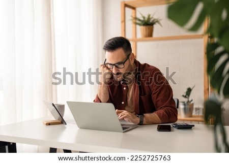 Focused young businessman using laptop working online in a home office. Professional businessman looking at laptop checking web market, e-learning webinar, having remote hybrid call sitting indoors.