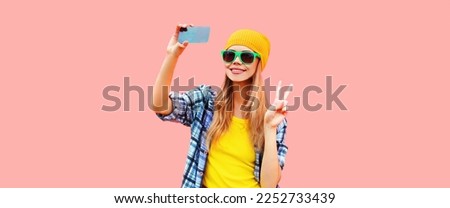 Portrait of modern blonde happy smiling young woman taking selfie with smartphone wearing colorful hat on pink background