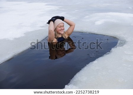 Winter swimming. Woman in frozen lake ice hole. Swimmers wellness in icy water. How to swim in cold water. Beautiful young female in zen meditation. Gray hat and gloves swimming clothes. Nature lake Royalty-Free Stock Photo #2252738219