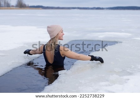 Winter swimming. Woman in frozen lake ice hole. Swimmers wellness in icy water. How to swim in cold water. Beautiful young female in zen meditation. Gray hat and gloves swimming clothes. Nature lake Royalty-Free Stock Photo #2252738229