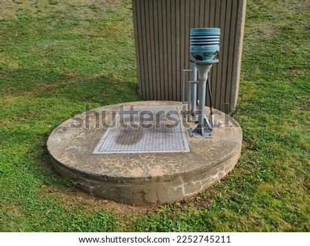 The top of a sewer lift located in Nova Scotia. There are two pipes. One is curved and looks like a cane. The other is capped with a light blue top. The cover is cement and round with a door hatch. Royalty-Free Stock Photo #2252745211