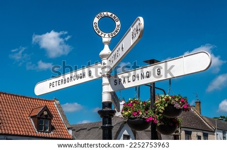 Old fashioned Holland County Council road sign in Crowland, Lincolnshire showing Peterborough, Spalding and Thorney. Also showing a thatched roof cottage, bright hanging baskets and blue sky.