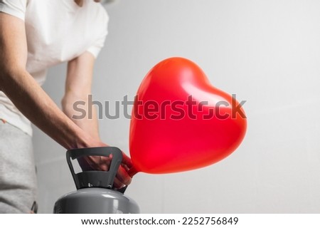 Hands of a young caucasian man inflate a red heart balloon with a helium compressor on a white background,side view close-up.Valentine's day concept,preparation,holidays.