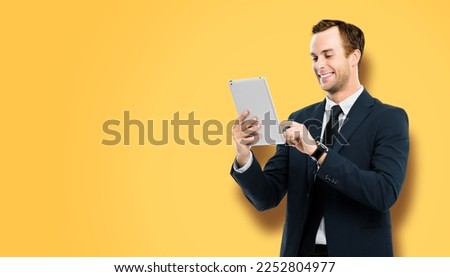 Portrait of smiling businessman in black suit using touchpad tablet pc, on vivid yellow background. Success in business concept studio picture. Copy space for ad text.