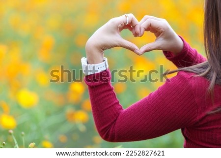 Blurred background,Young woman wearing red dress raises her hands to make heart symbol to represent love, friendship and kindness on background of field of yellow flowers. heart symbol, Love concept