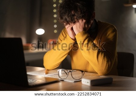 Tired and exhausted young man hides eyes with hands at laptop pc late in the evening. Depressed male student sitting in front of computer screen at night. Concept of anxiety. Copy space Royalty-Free Stock Photo #2252834071