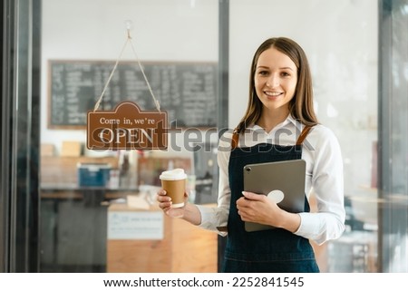 Young friendly female pretty caucasian coffee shop woman owner in apron with open sign, small business owners smiling and service take away orders from food delivery application on tablet.