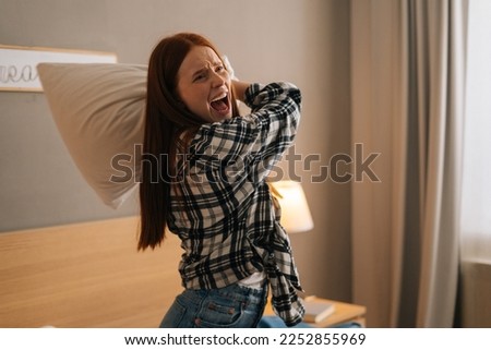 Portrait of angry annoyed woman with open mouth shouting and yelling with aggressive expression, throwing pillow sitting on bed in bedroom. Furious crazy redhead female vented anger in privacy of home Royalty-Free Stock Photo #2252855969