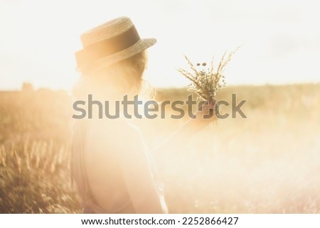 Close up girl in straw hat with bouquet surrounded by sunlight glare concept photo. Side view photography with meadow on background. High quality picture for wallpaper, travel blog, magazine, article