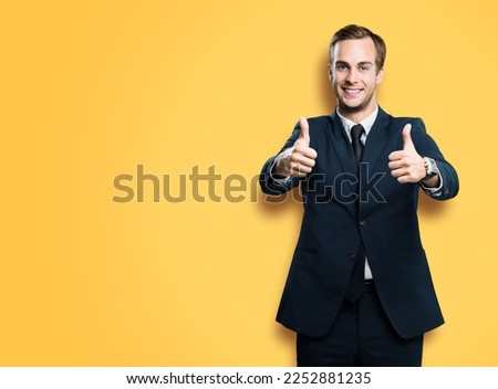 Cheerful smiling businessman in black confident suit showing two thumbs up like hand gesture, on vivid yellow background with copy space for ad, slogan or text. Successful business man.