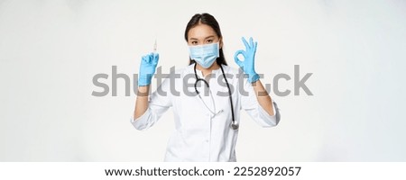 Covid-19 vaccination and healthcare. Asian woman doctor in medical face mask and gloves, holding vaccine syringe and show okay sign, white background.