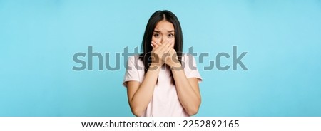Shocked asian woman covers mouth screams, gasping startled, scared of smth, shuts her lips, standing over blue background. Royalty-Free Stock Photo #2252892165
