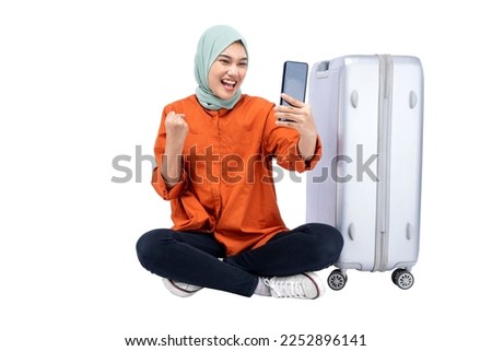 Asian Muslim woman in a headscarf sitting with a suitcase while using a mobile phone isolated over white background