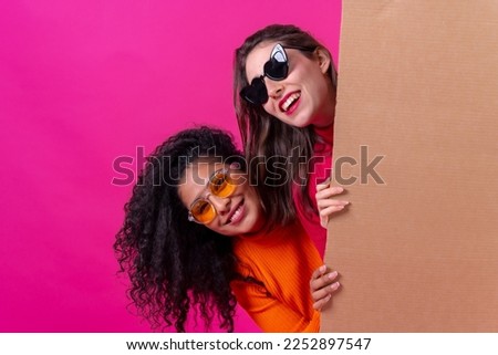 Two smiling beautiful girl friends hold a cardboard sign on a pink background