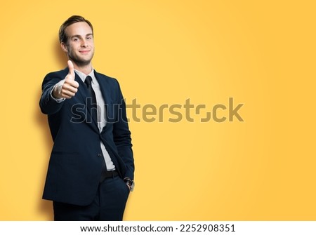 Smiling businessman showing thumb up like sign gesture, in black suit, on vivid yellow background. Confident business man. Copy space for slogan or text.