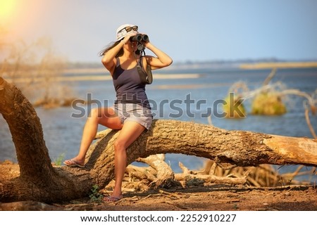 On safari in Zimbabwe: Fit Woman sitting on tree trunk over Zambezi river observing African Nature through binoculars. Independent woman on safari trip. Mana Pools National Park, Zimbabwe, Africa. Royalty-Free Stock Photo #2252910227
