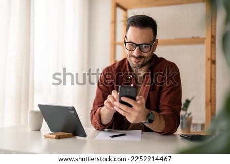 Happy smiling businessman wearing casual clothes and using modern smartphone in his home office during the day, typing, touching the screen, browsing the internet or writing text messages. Royalty-Free Stock Photo #2252914467