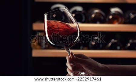 Close up female hand swirling red wine in wine glass. Wine expert tasting, rating and drinking wine, bottles in background. Royalty-Free Stock Photo #2252917461