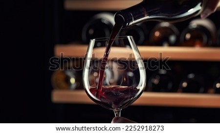 Close up female hand pouring red wine to glass. Wine expert, sommelier tasting, rating and drinking wine, bottles in background. Restaurant, wine bar, waitress.