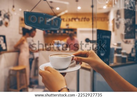 Coffee cup latte art in women's hands in background of interior of coffee shop. Local small businesses at food service.