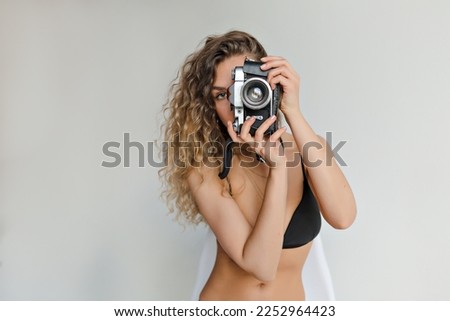 Close up indoor portrait of appealing stylish woman with wavy long light hair wearing black swim suit is making photo on retro camera over isolated background 