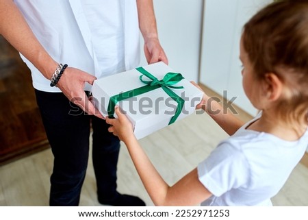 Little girl is giving her handsome father a gift box on Father's day, unrecognizable daughter congratulating dad and giving present on birthday at home. I love you, dad. Happy Father's Day.
